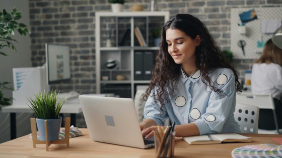Woman working at a desk with a laptop, pens, and books, surrounded by plants and shelves.