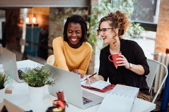 Two women with laptops and a coffee mug sitting at a table in an office.