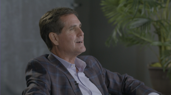 A man in a suit is sitting on a chair with a potted plant behind him.