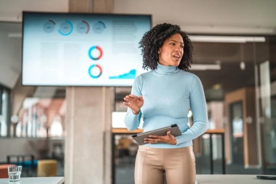 Woman in a turtleneck top standing in front of a screen giving a presentation at a meeting.