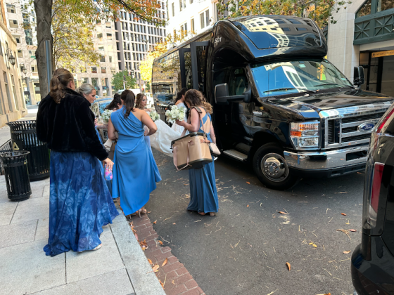 A group of bridesmaids stand on the sidewalk outside of a black limo waiting for the bride.