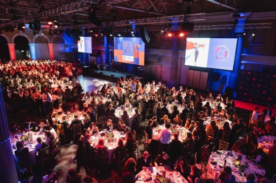 A large room with many people sitting at tables with a large screen on the wall and a stage.