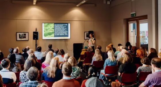 A woman stands on stage, speaking to a group of people, with a green screen behind her.