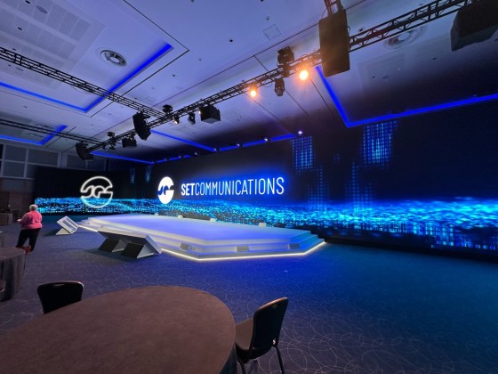 A conference room with a large screen showing the words "SET COMMUNICATIONS" and a person standing nearby.