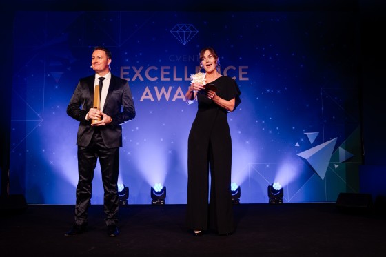 A man and a woman standing on stage holding trophies and a flower in their hands.