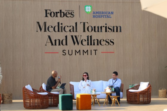 Three people sit in chairs on a stage with a Forbes Medical Tourism and Wellness Summit banner.