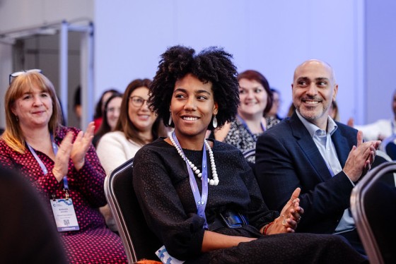 Smiling audience at an event with a purple wall and white doors and a metal railing in the background.