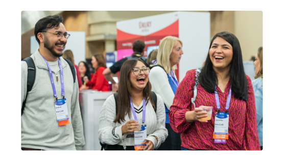 Three people are walking together at a conference or event, all smiling and appearing to enjoy themselves. Each person is wearing a name badge and a purple lanyard, and two of them are holding drinks. The background shows other attendees and a booth area with signage, while the overall atmosphere is lively and welcoming.