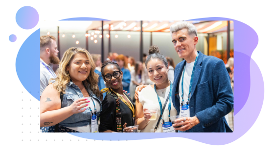 A photo of four people standing together at an event, smiling at the camera and holding drinks, with name badges and lanyards visible. The background features a softly lit indoor setting with other attendees, and the image is framed by a blue and purple abstract shape with circular accents