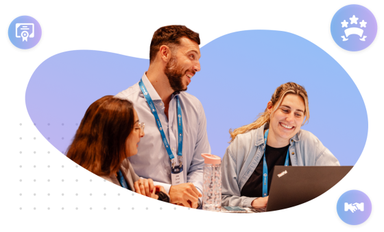 Three people are sitting together at a table, engaged in a collaborative discussion. Two women and one man are visible, all wearing blue lanyards with name badges. The man, standing or leaning forward, is smiling and looking at one of the women, who is also smiling while working on a Lenovo ThinkPad laptop. The other woman, with long brown hair and glasses, is listening attentively. A clear water bottle with a pink lid is on the table. The group is set against a blue, abstract, rounded background shape. 