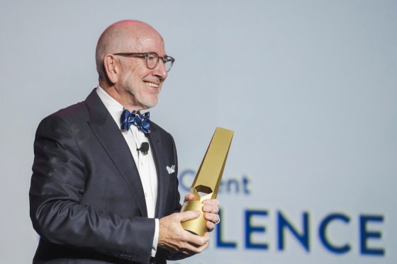Man presenting on stage in a tux with gold award