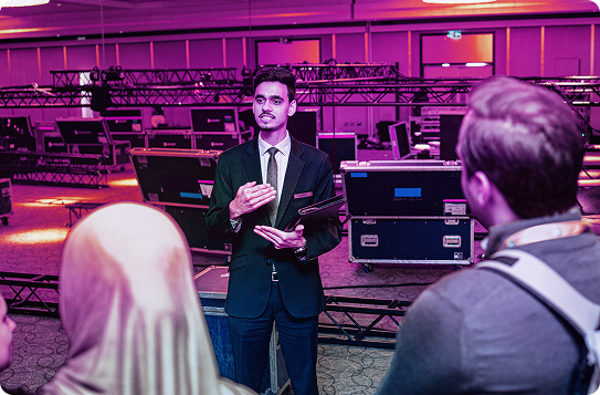 A venue staff member in a suit speaks to a small group during an event setup, with equipment cases, trusses, and staging materials behind him in a large ballroom.