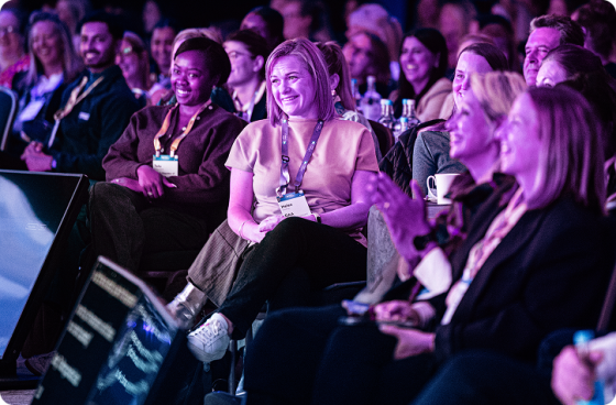 Audience members seated in a conference hall smiling and laughing during a presentation, with large display screens visible at the front.