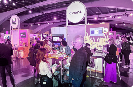 Cvent booth at a busy event expo, with attendees interacting and displays of branded tote bags, pins, stickers, and other giveaway items arranged on shelves and carts.