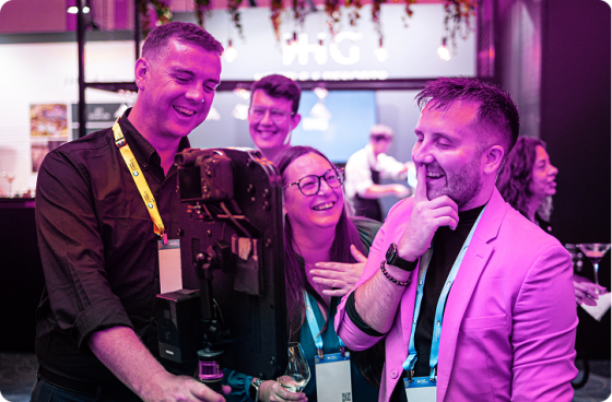 A group of conference attendees laugh together while watching a recording or photo on a camera setup during a networking event.