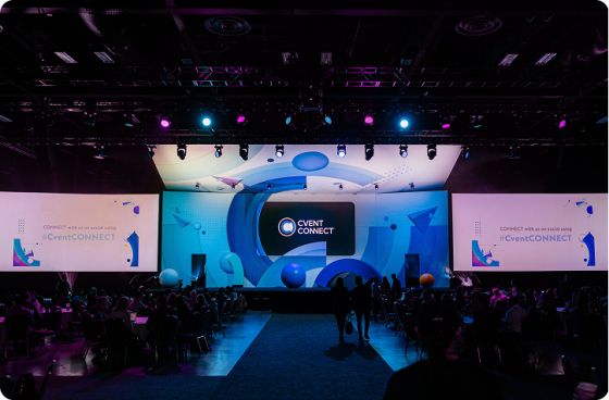 A large conference hall with a wide, brightly lit stage displaying the Cvent CONNECT logo and blue graphic visuals, surrounded by large projection screens and an audience seated at round tables in a dimly lit room.