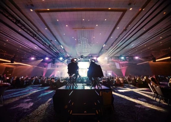 Two camera operators film a glowing stage in a vast ballroom filled with seated attendees under dramatic purple event lighting.