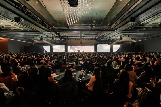 A massive ballroom audience seated at round tables watches a cinematic presentation on a wide panoramic stage screen.