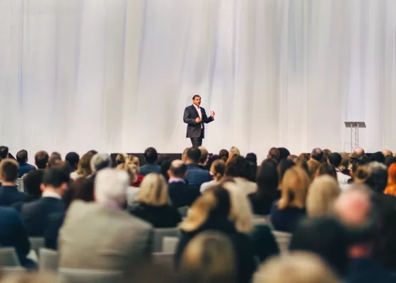 A keynote speaker gestures on stage before a large seated audience in a bright, draped conference hall.