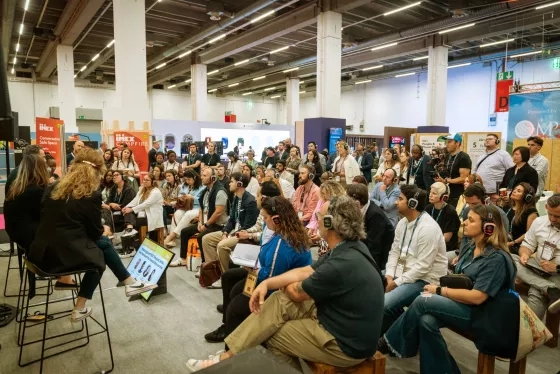 Wide view of a seated crowd wearing wireless headphones listening to a panel session at a trade show.