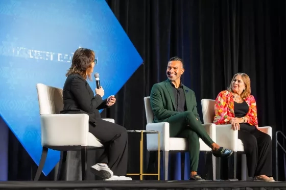 Three panelists seated on stage, one speaking into a microphone during a discussion.