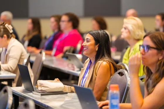 Woman smiling and looking up during a conference session, seated at a table with a laptop.
