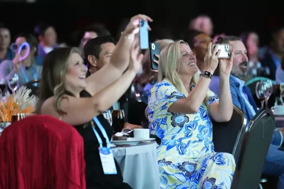 Two women raising phones to take photos during an event, smiling and cheering.