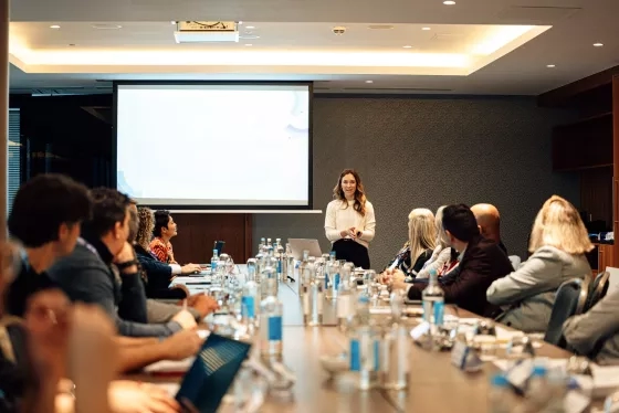 Woman presents to a seated audience at a long conference table with water bottles.