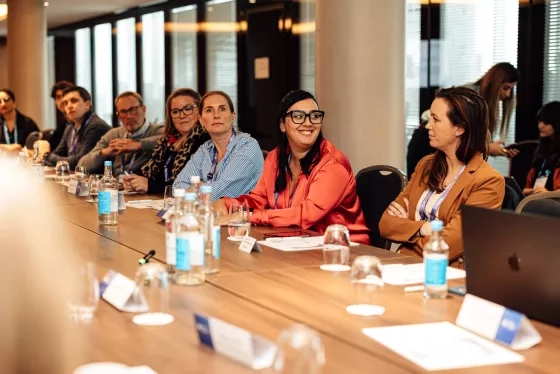 Attendees at a conference table listen to a presentation, one woman smiling.