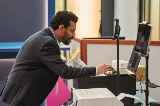 Man leaning over a laptop and printer setup with a smartphone on a mount, working at a tech station.