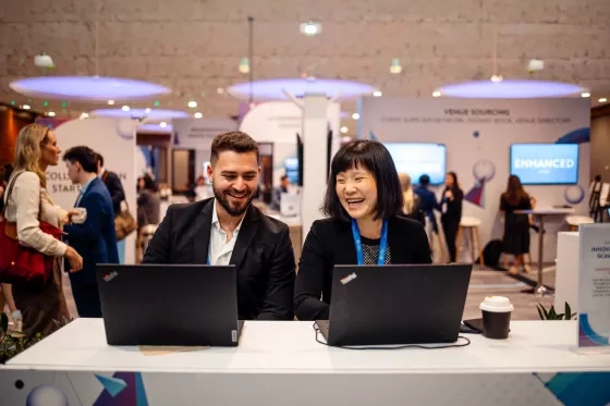 Two colleagues laughing together while working on laptops at a conference expo booth.