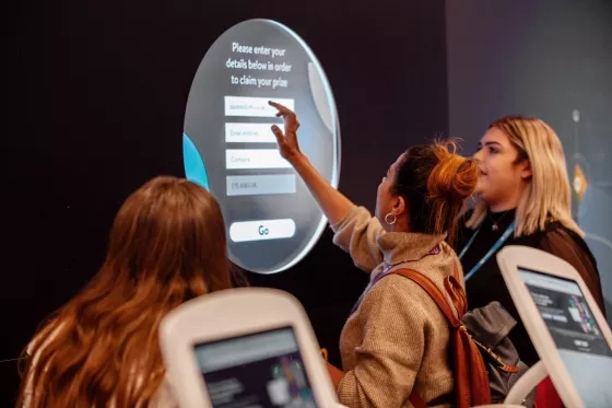 Woman reaching up to a large touchscreen kiosk displaying a form to claim a prize.