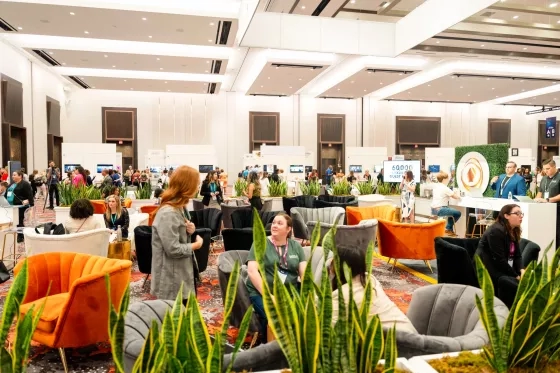Wide view of a conference lounge area with orange and gray chairs, plants, and attendees mingling.