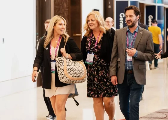 Three conference attendees wearing lanyards walking and conversing in a bright event hall with sponsor booths.