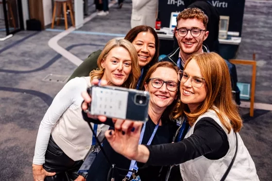 Five conference attendees with lanyards smiling and taking a group selfie together indoors.