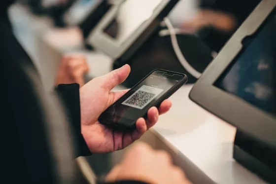 Hand holding a smartphone displaying a QR code near event registration kiosks.