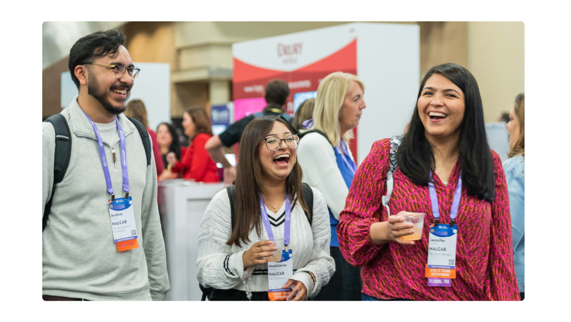 Three people are walking together at a conference or event, all smiling and appearing to enjoy themselves. Each person is wearing a name badge and a purple lanyard, and two of them are holding drinks. The background shows other attendees and a booth area with signage, while the overall atmosphere is lively and welcoming.