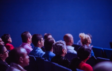Several people are sitting in chairs in a theater, watching something on a screen.