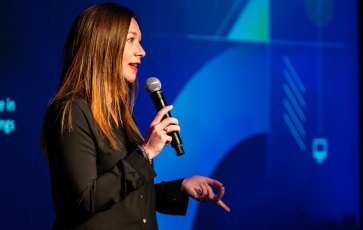 A woman wearing a black shirt and holding a microphone is speaking in front of a blue screen.