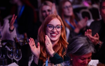 Smiling young woman claps at an event with friends, surrounded by glasses and bottles on a table.