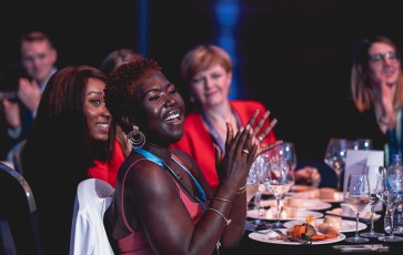 A woman at a dinner table with other people in a room, clapping her hands and smiling.