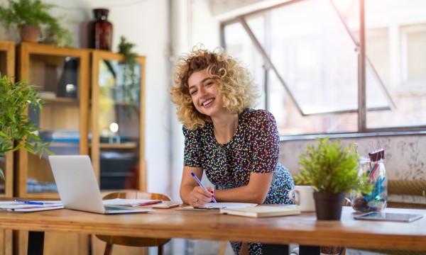 A smiling woman sitting at a desk with a laptop, pen, paper, and potted plants.