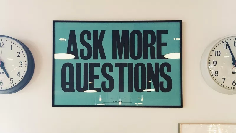 A wall with two clocks and a framed sign that says "Ask more questions" in black letters.